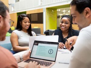 Four social work students sit around a desk smiling. Two face the camera on the other side of the desk, both looking at a laptop in front of them. Two students are facing away from the camera and looking at a laptop in front of them.