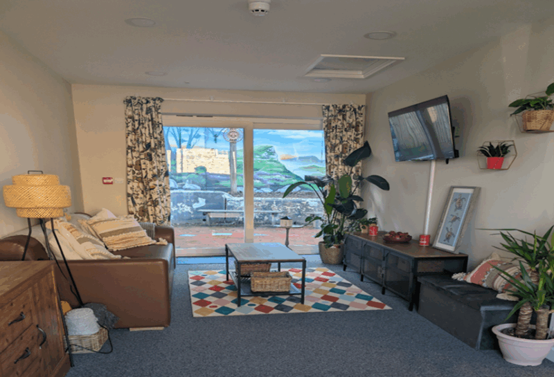 The lounge area which has large gladd patio doors out to teh garden, leather sofas, a colourful rug under a coffee table, and a TV on the wall.