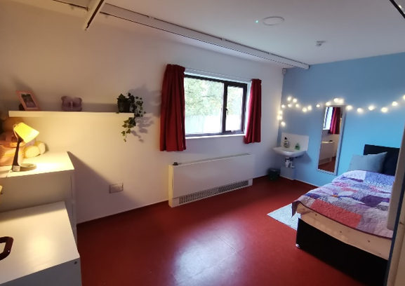 A bedroom with a white wall, and a blue wall with string lights on it, red floor and curtains, and a bed with a colourful cover on it.