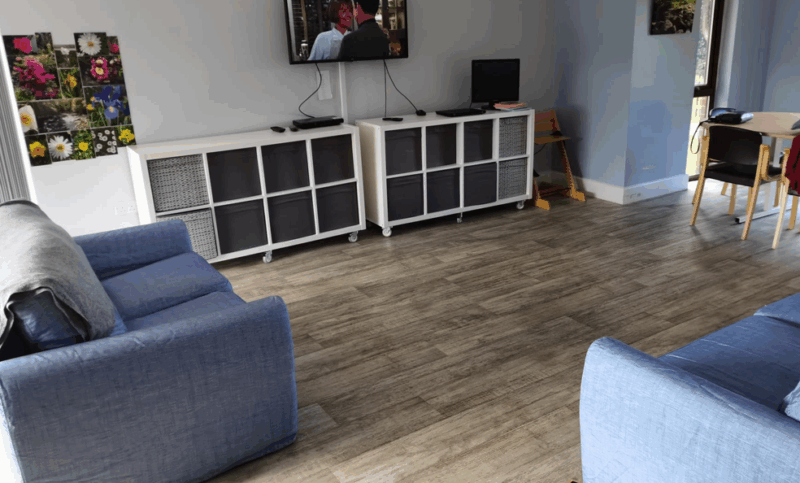 The lunge area with a sofa and armchair, storage units, and a TV on the wall, with a table and chairs at one end.