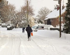 road covered in snow with person walking down the road, a person in the background is shoveling snow from their drive