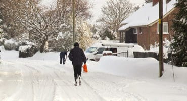 road covered in snow with person walking down the road, a person in the background is shoveling snow from their drive