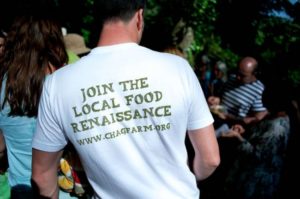 group of people gathered, man stood with back to camera wearing a white t-shirt that says join the local food renaissance www.chagfarm.org