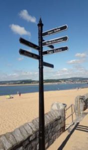 Black multi directional fingerpost sign on pavement next to the sandy beach at Exmouth