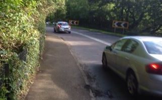 cars on road with pavement and hedges running alongside coming up to a sharp left hand bend with the black and white roadsigns that symbolise this