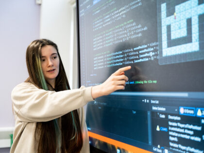 A young woman at South Devon College with brown and green hair is pointing at a large screen with code on it.