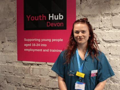a young woman with red curly hair is standing in front of the Youth Hub sign in scrubs.
