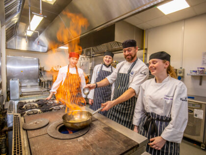 Apprentices from South Devon College (two men and a woman) in chef's whites and pin stripe aprons work with a chef who is an older man with a short beard. They are in a catering kitchen and there is a pan with a dramatic flame from in.