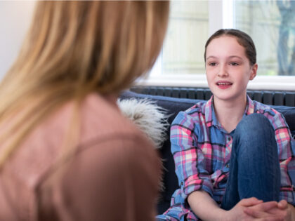 A young woman with a blue and pink check shirt is sat on a sofa with her knee pulled up against her chest, she is having a conversation with an older woman.