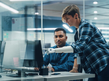 A woman with short cropped hair and a prosthetic am is showing an employee something on a computer screen in an office.