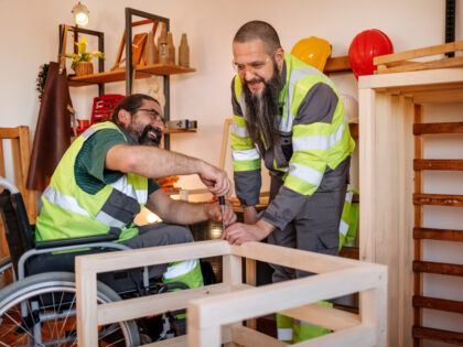 Twi men with big beards are working in a carpentry workshop, they are assembling a chair, the man in the wheelchair is helping another man. They are both wearing high vis jackets.