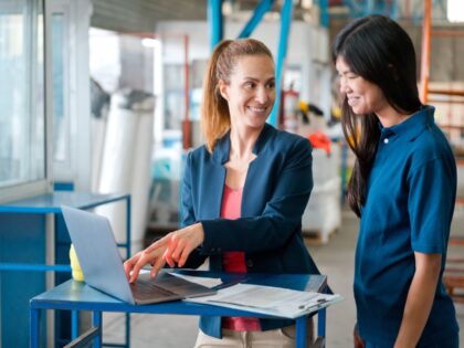Female manager wearing navy blue jacket standing in warehouse, using laptop and talking with employee.