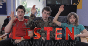 The image shows three young people, who are members of our Champions for Change group, sat down and holding their arms up with their flat palms facing forwards.  The image is accompanied by the word ‘Listen’ in capital red text.