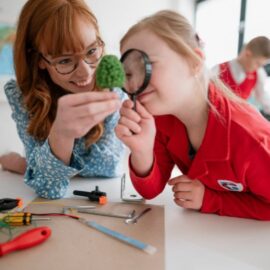 A young girl sitting at a table with a woman. The girl is looking through a magnifying glass