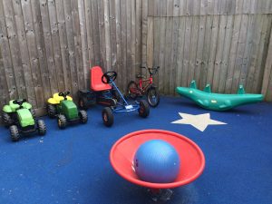 A photograph of some of the toys in the outdoor play area in the garden at Barnes: two little tractors, a bicycle, a go-cart, a large blue rubber ball and a see-saw.