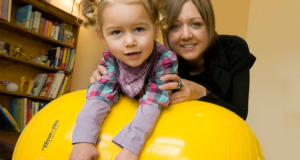 Mother holds the arms of her child who is balancing on a yellow exercise ball