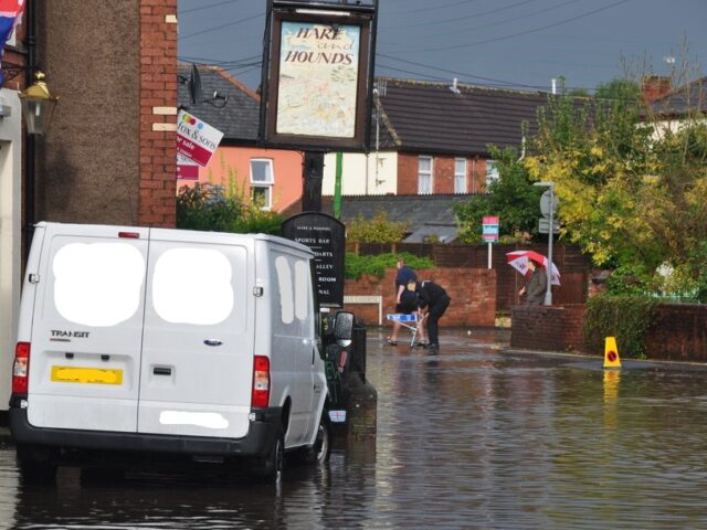 Chapel Street, Tiverton, Copyright Lewis Clarke. Street with flooded road surrounded by houses. Pub and white van in foreground. People in background. https://www.geograph.org.uk/photo/3174972