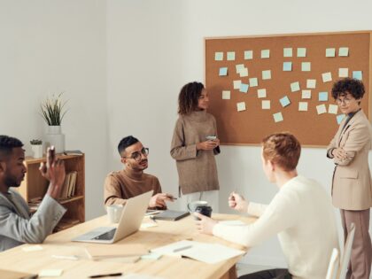 Two women standing on either side of a corkboard covered in sticky notes. One of the women is looking at three other people who are sitting around a table having a discussion