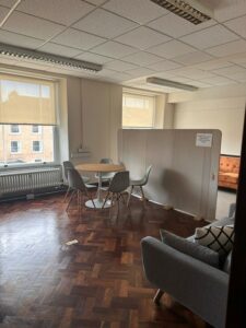 A picture of the room, it is bright and spacious. There are two windows with cream coloured roller-blinds. The first part of the room has a varnished wooden floor. There is seating in the room - tables and a round table, and grey sofa and cushions. There is a grey space divider to the right and beyond the divider, the room has a beige carpet and an orange sofa.Please remove your shoes before walking on the carpet.