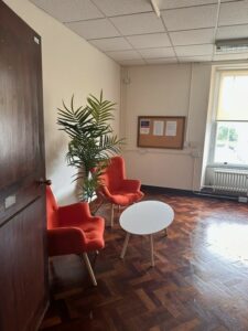 The room to the door is open and just inside there are two bright orange chairs with armrests, a large pot plant, and a white oval table.