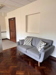 A grey sofa is also available in the room. There are two white cushions on the sofa with black lines as a pattern. There is a door at the far end but this is locked shut. Behind the sofa is a small recess.