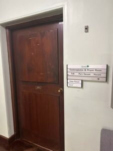 A sign to the right of the door says Devon County Council Contemplation and Prayer Room. There is a sliding arrow underneath which can indicate if the room is full (slide left), part vacant (slide to the middle) or vacant (slide to the right).