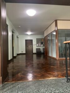 Picture of the corridor leading to the contemplation and prayer room. The floor is dark wooden parquet flooring. The wooden door at the end is dark brown. To the right there is a glass enclosed meeting room.