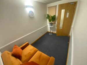 This picture is looking at the room from the angle of the orange sofa. The door at the end of the room is opened slightly. The room is small and oblong shaped, it is simply furnished and has a sense of calm.