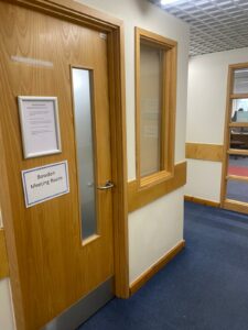 A corridor with a navy blue carpet and a wooden door with a notice on it saying Bowden Meeting Room. To the right of the door is a window.