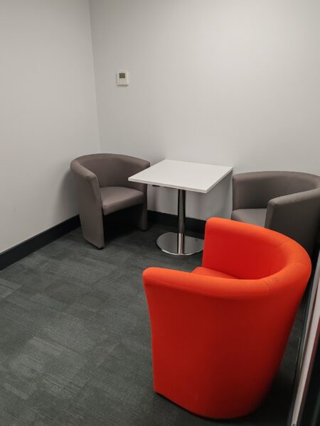 Appledoor Room, Taw View. Picture of inside the room. The walls are light grey, the carpet is a dark grey colour. There are two grey comfy armchairs around a small white table, and an orange comfy armchair.