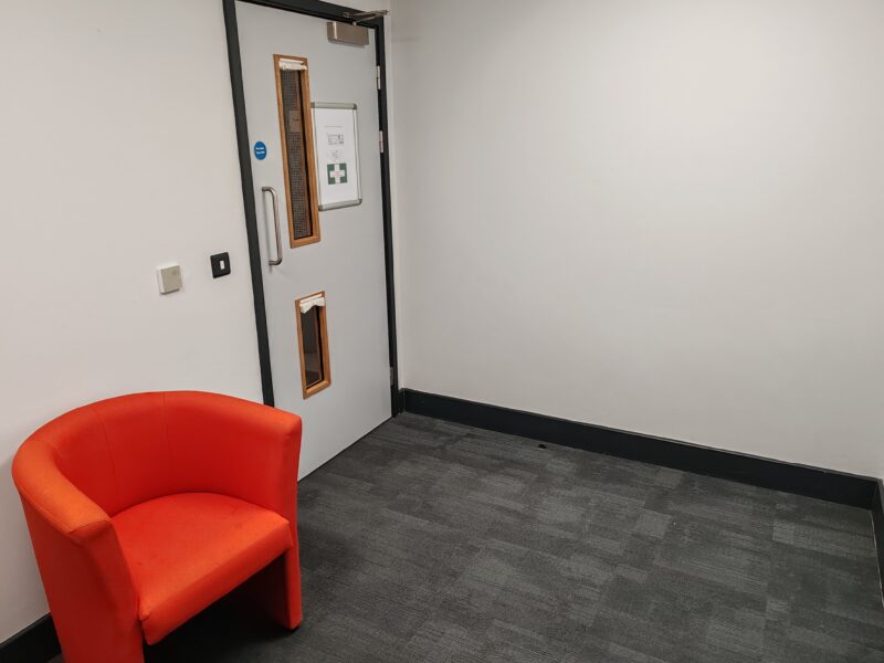Appledoor Room, Taw View. Picture from inside the room showing the inside of the door which has two narrow windows and blinds for privacy. There is a dark grey carpet and orange comfy chair beside the door. The walls are plain and pale grey or white in colour. There is a first aid notice on the door.