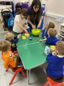 A group of children sit around a table whilst a childcare practitioner mixes something in a bowl.