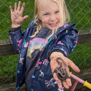 A young girl is outside with her coat on. In her outstretched hand is a worm.