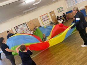 Children in a hall playing with a colourful parachute canopy.