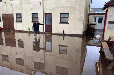 A photograph of a white house surrounded by flood water with a man wading through.