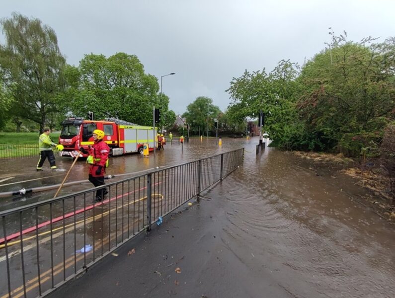 Flood water on a road with a parked fire engine and firefighters working to clear the water