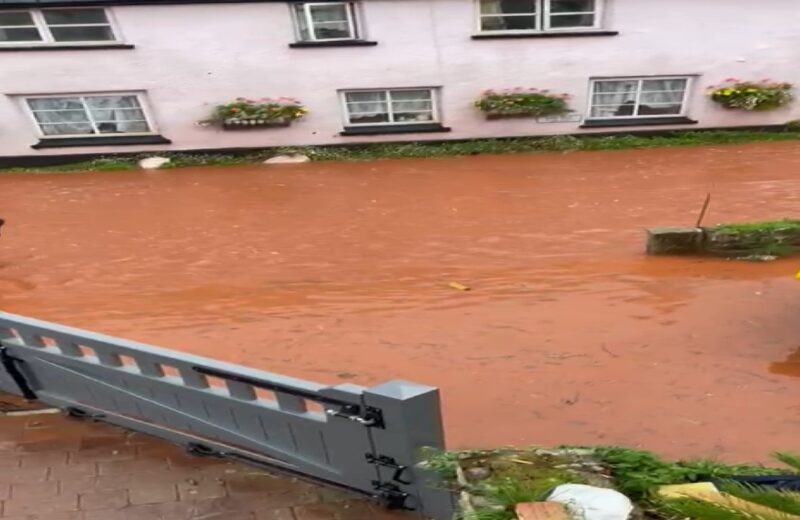 Muddy flood water flowing down a road with a closed flood gate in the foreground, there is no flood water on the property side of the flood gate