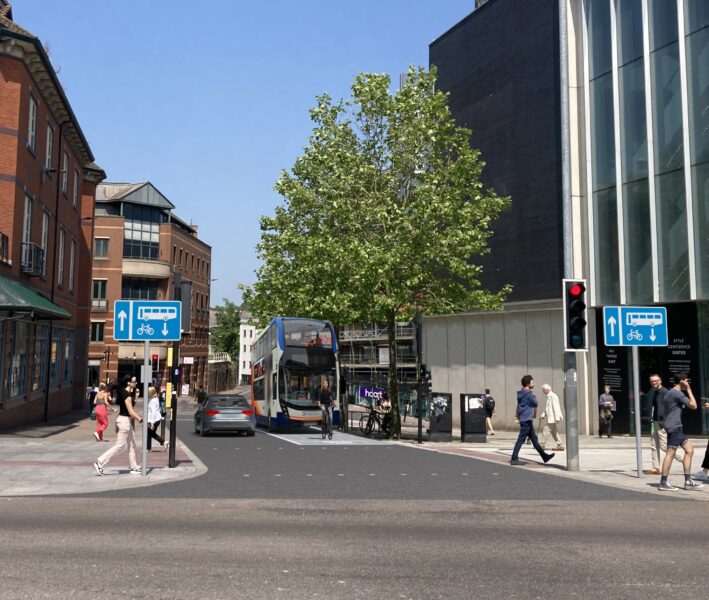 : A visualisation of the proposed New North Road bus gate and bus lane between Waterstones and John Lewis looking towards New North Road. The image shows the new contraflow bus and cycle lane signage on either side of the carriageway as well as the new road surface including the block effect imprint on the bus lane. The image shows a double decker bus and passenger car passing side by side with a cyclist in front of the bus. 