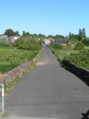 A colour photograph of a road bounded by low stone walls, raised above the level of the surrounding low-lying grassland. The buildings of a setttlement are visible along the road in the distance