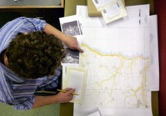 A colour photograph, taken from above, showing an aerial photograph interpreter seated at a desk, locating the position of 1940s black and white photographs on a modern map.