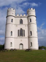 A colour photograph of a tall crenelated structure, circular turrets at each corner, rendered in white-grey with numerous arched window openings and mown grass and picnic tables in front