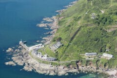 A colour oblique aerial photograph of the rocky coastline south of Plymouth, with tiers of low buildings on the slopes down to the shore. A substantial earthwork ditch is visible to one side, perpendicular to the contour