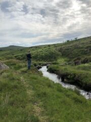 A colour ground photograph of a moorland landscape with a curving water channel in the foreground and a person stood next to it.