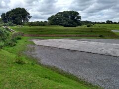 A colour ground photograph taken from one arm of a dispersal pen. The concrete area of hardstanding between the earthworks and an entrance into the embanked bunker are visible.