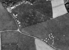 A black and white aerial photograph, in which hut bases are visible as pale rectangular features, a zig-zag trench is indistinctly visible as an earthwork along the road, and pale areas of compacted ground, or parchmarks, on the location of the rocket projectors, are visible in several adjacent fields.