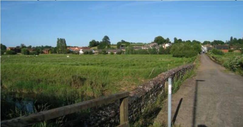 A colour photograph of a road that runs over a bridge with low stone parapet, and is raised above the level of the surrounding low-lying grassland. The buildings of a setttlement are visible on the horizon.