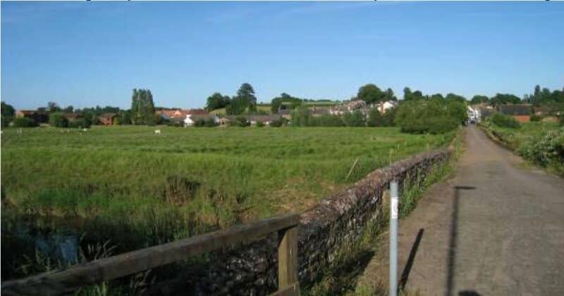 A colour photograph of a road that runs over a bridge with low stone parapet, and is raised above the level of the surrounding low-lying grassland. The buildings of a setttlement are visible on the horizon. 