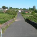 A colour photograph of a road bounded by low stone walls, raised above the level of the surrounding low-lying grassland. The buildings of a setttlement are visible along the road in the distance