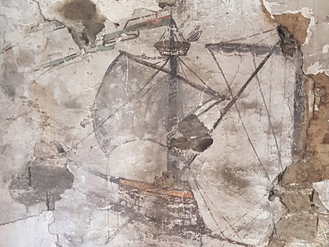 A old mural of a warship on a wall where some plaster is flaking off