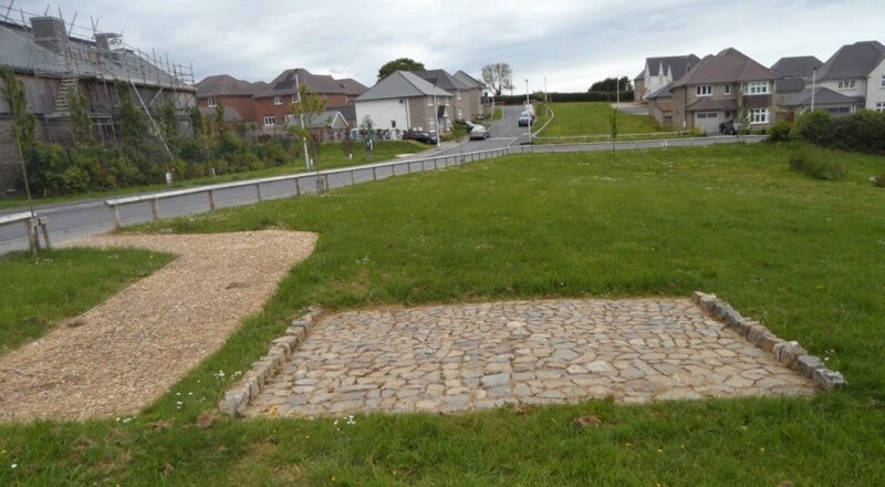 A colour photograph of a residential development, with a grassed area in the foreground, within which sits a section of a slightly convex roadway formed of irregularly-shaped closely-laid stones with a narrow stone kerb on either side.
